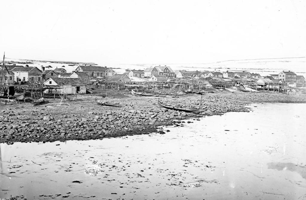 Vadsø på slutten av 1800-tallet. Trehusbebyggelse ved sjøen. Noen få båter i forgrunnen. Foto.