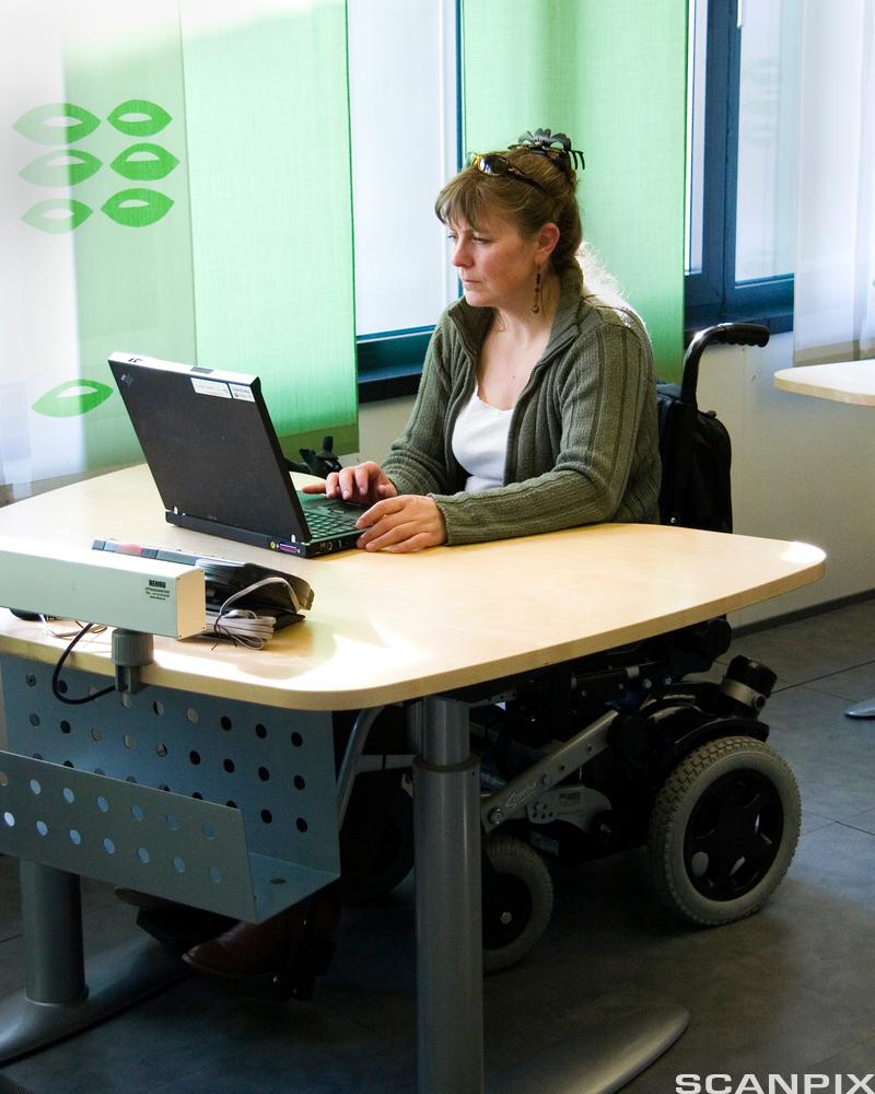 Ei kvinne i rullestol sit ved ein kontorpult. Foto
