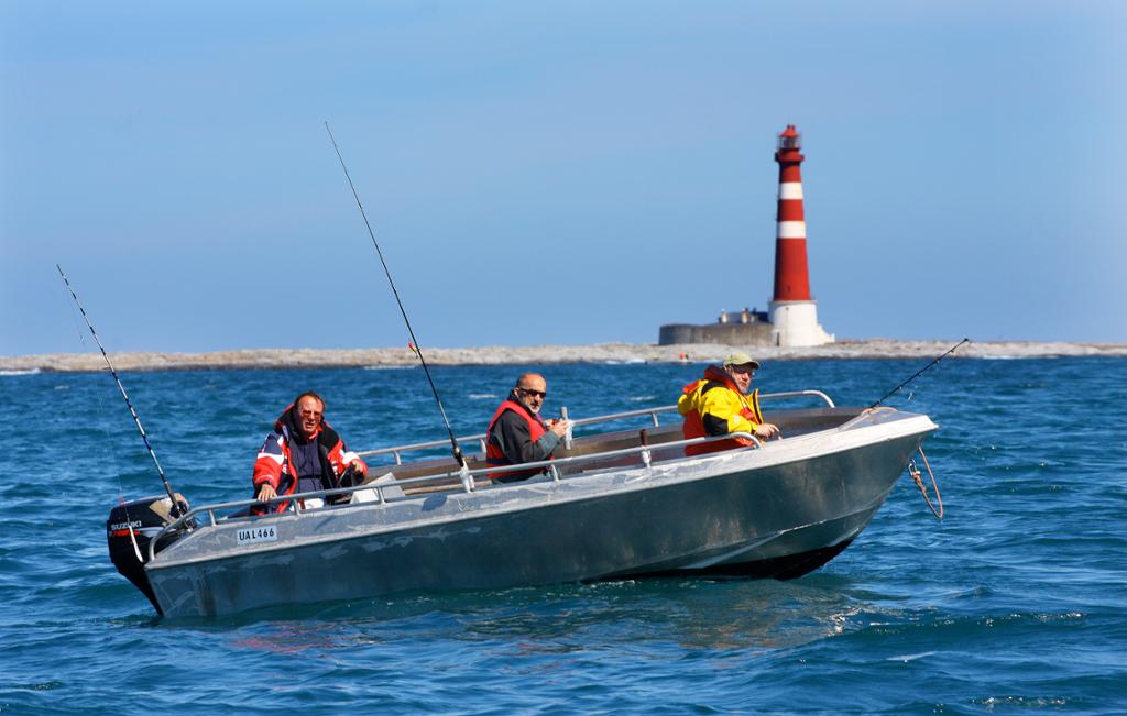 Tre fritidsfiskarar i ein liten aluminiumsbåt med eit fyrtårn i bakgrunnen. Foto.