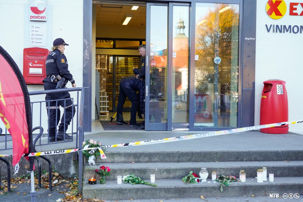 Inngangspartiet til en Extra-butikk, med sperrebånd og politi som arbeider på stedet. På trappen foran inngangsdøra er det  blomster og lys. Foto.