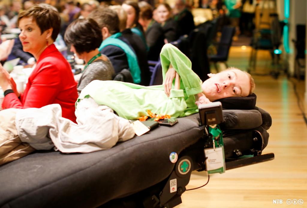 Funksjonshemmet ung mann ligger på en seng som styres av elektronikk, i en forsamling med mange andre mennesker. Foto.