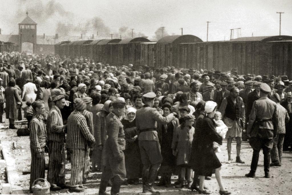 Jødene sorteres etter ankomst i Auschwitz. Stor folkemasse med menn i uniform og menn, kvinner og barn. Togsett og hovedinngangen i bakgrunnen. Foto.