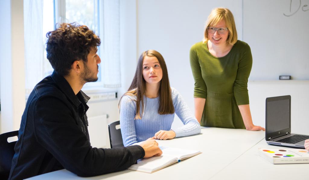 En kvinnelig lærer står ved et skrivebord og smiler. Hun er i samtale med to elever – en gutt og en jente. Foto.