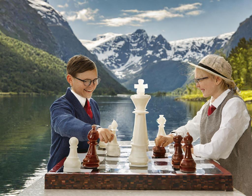 To personer sitter ved et sjakkbrett med store sjakkbrikker. Brikkene har ikke samme størrelse. Fjell og fjord i bakgrunnen. KI-foto.