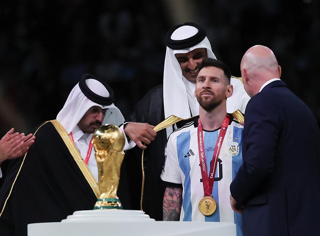 Ein mann i argentinsk fotballdrakt med gullmedalje på brystet får på seg ei kappe av to menn som er arabisk kledde og ein mann som er vestleg kledd. Foto. 