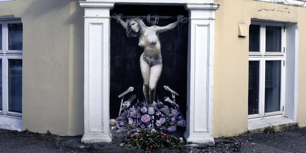 Veggmaleri som viser en kvinnelig politiker som er korfestet. Foran føttene mange mikrofoner og blomster.Maleri.