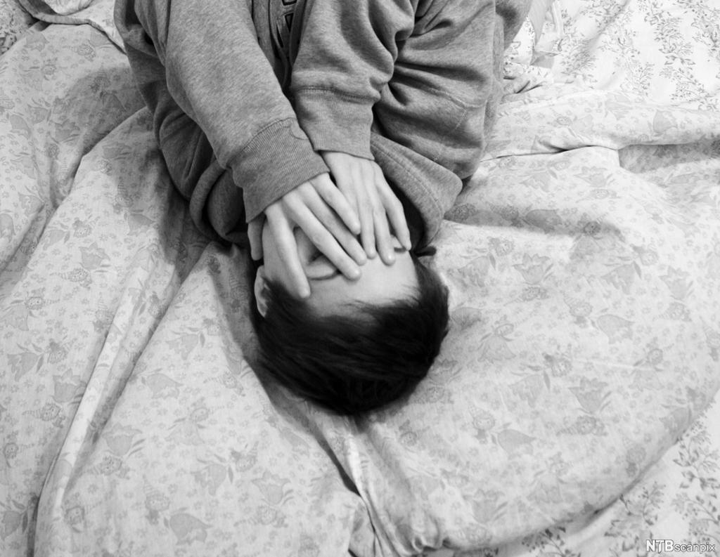 Ung kvinne ligger på senga med hendene over ansiktet. Svart-hvitt-foto. 