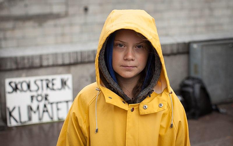 Greta Thunberg, wearing a yellow raincoat. A poster behind her reads School Strike for the Climate in Swedish. 