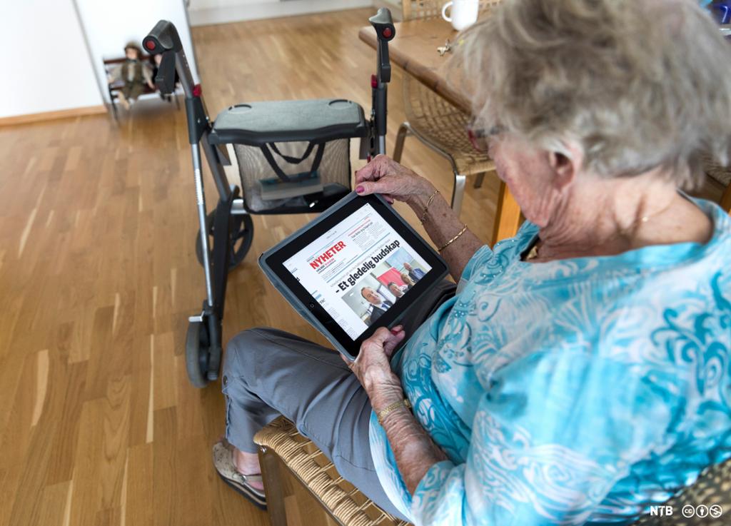 Eldre dame med rullator leser avis på nettbrett. Foto.