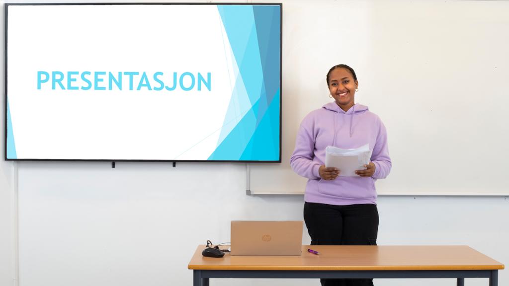 Ei smilende jente foran en stor skjerm med teksten Presentasjon. Hun holder et manuskript i hendene. Foto.
