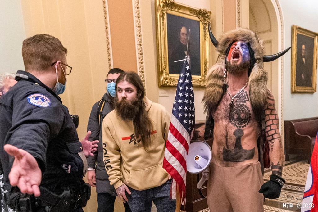 Photo: A man in uniform is facing a group of men in a corridor. One of the men wears a hat with fur and horns; he has many tattoos. Another is wearing a yellow college sweater; he has long hair and a beard, he is carrying a flag. There are two more men obscured from view by the others in the picture. 