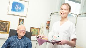 Kvinnelig helsefagarbeider står med matserveringsbrett. I bakgrunnen sitter en smilende eldre pasient i stolen. Foto.