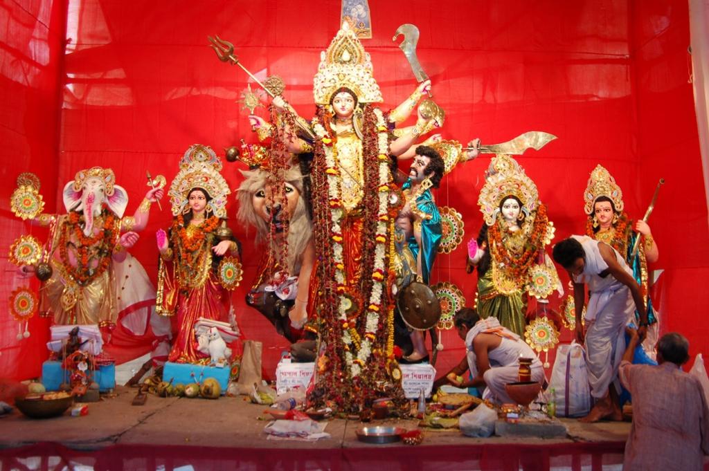 Fargerike statuar av gudar med ein stor kvinnestatue med mange armar i midten. Ein mann legg fram gåver ved føtene hennar. Foto.