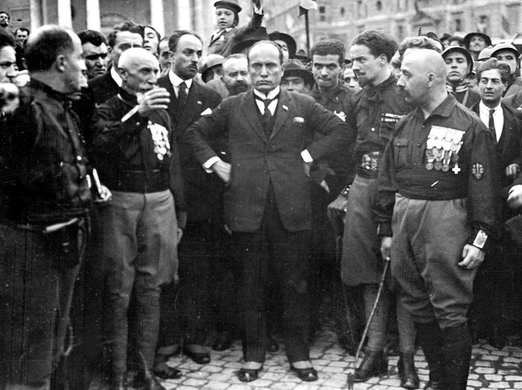 Benito Mussolini i dress midt i en stor mengde menn i svarte skjorter. Mennene nærmest Mussolini har medaljer på brystet. Fotografi. 