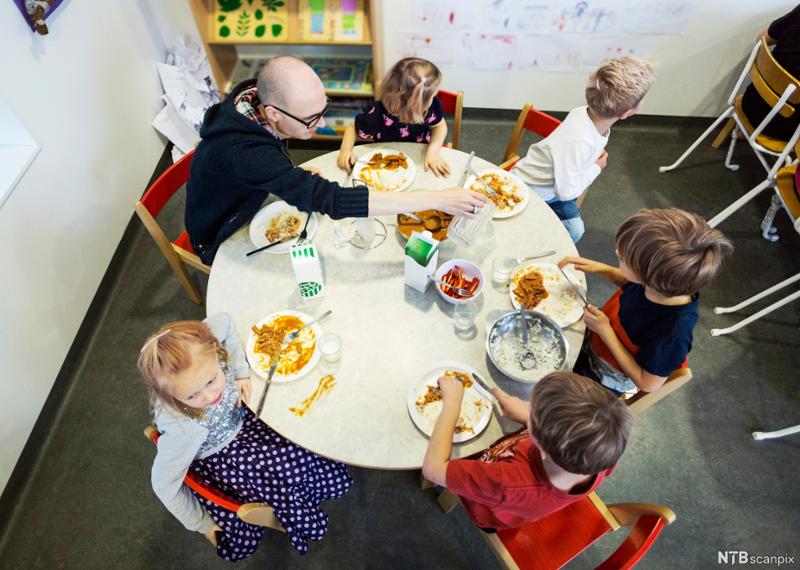 Barnehagebarn sitter rundt bordet sammen med en voksen. Foto. 