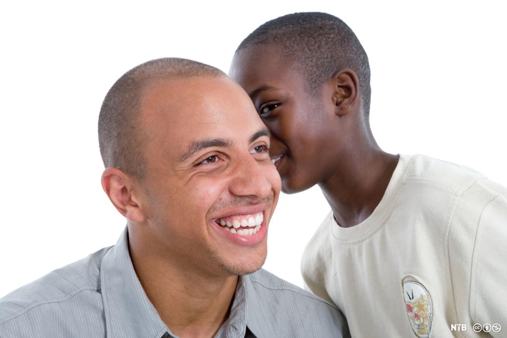 En ung gutt hvisker noe i øret til en mann. Begge smiler. Foto.