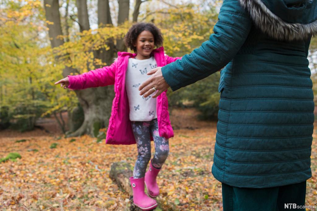 Ei smilende jente med rosa jakke og støvler balanserer på en tømmerstokk. En voksen strekker ut hendene for å ta imot henne. Foto. 