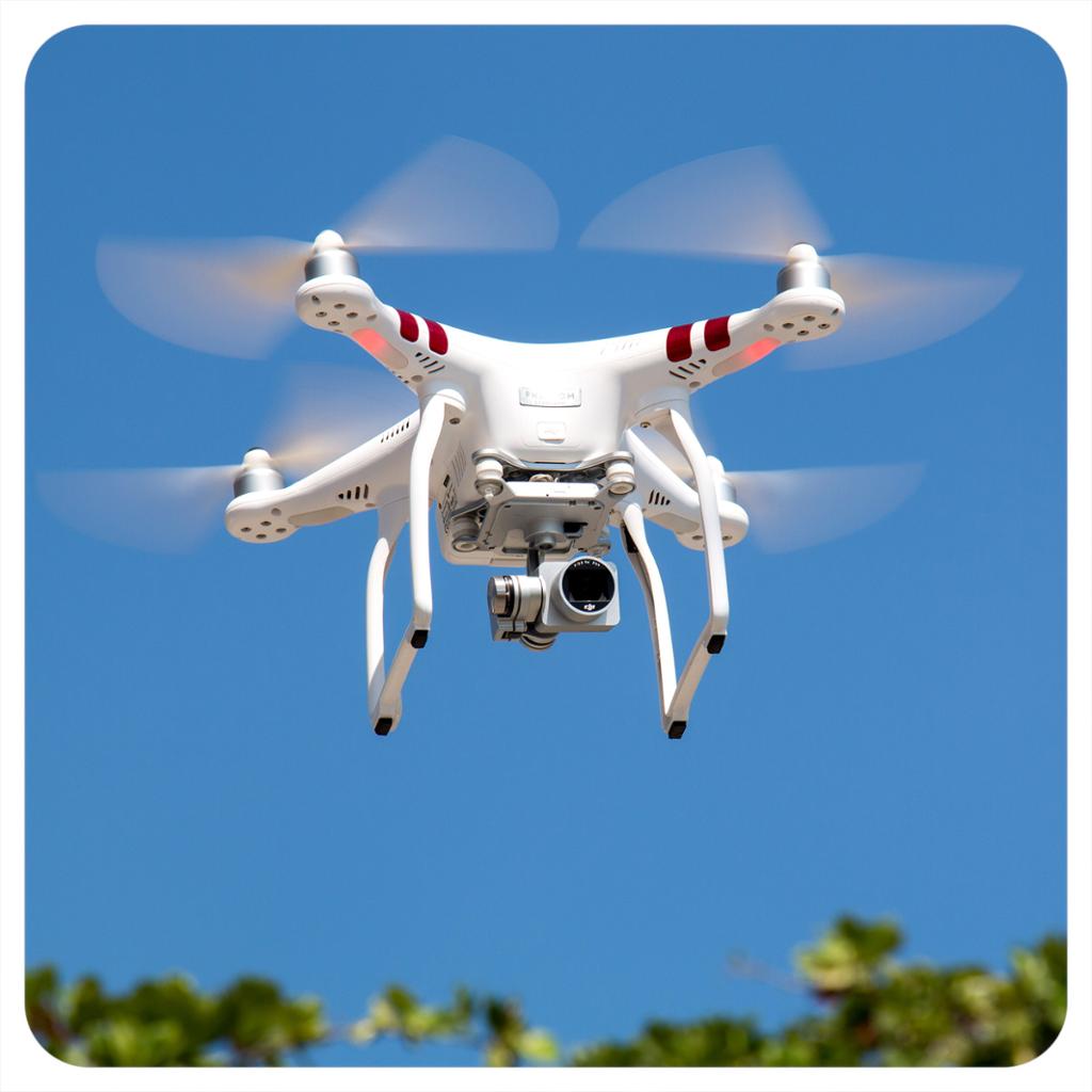 Hvit drone med fire propeller og påmontert kamera flyr i lufta. Det er blå himmel og bra vær. Foto.

