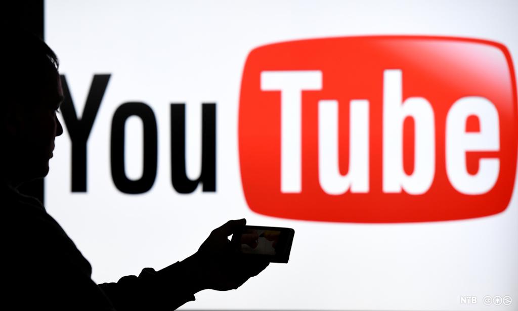 En gutt med mobiltelefon står foran logo av YouTube. Foto.