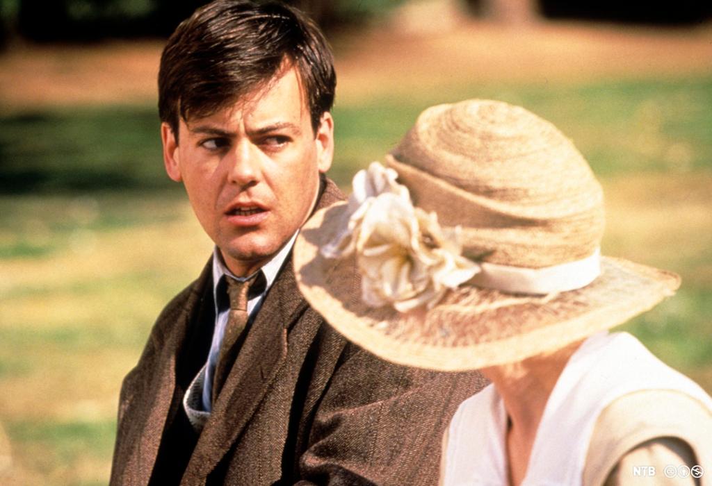 Photo: From the film Mrs Dalloway. We see Rupert Graves in the role of Septimus Smith. He is a young dark haired man, wearing a brown suit. A woman in a straw hat is looking at him. 