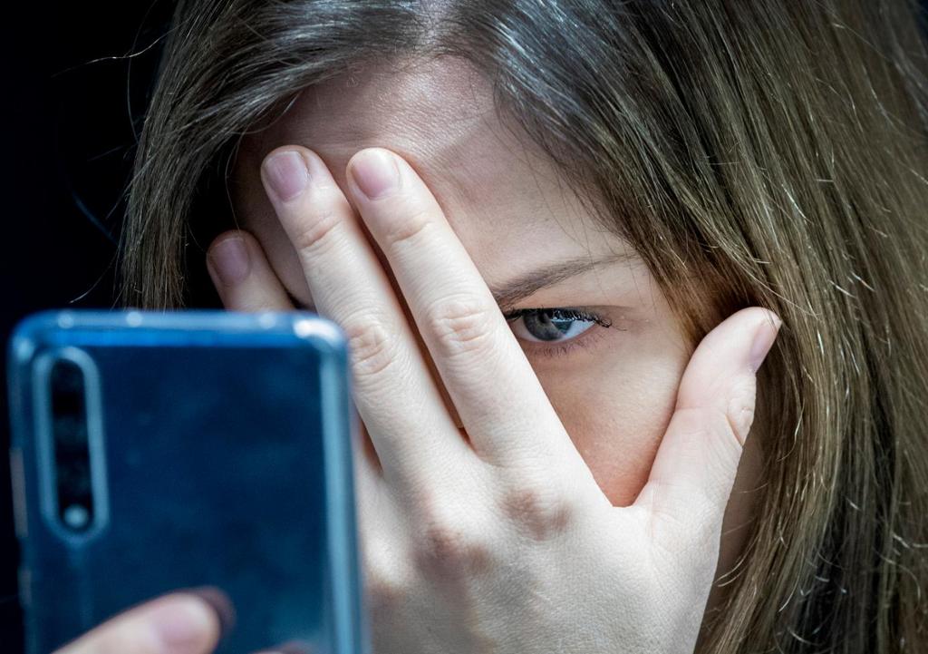 Kvinne leser hatefulle ytringer på mobiltelefon og holder hånden foran halve ansiktet. Foto.