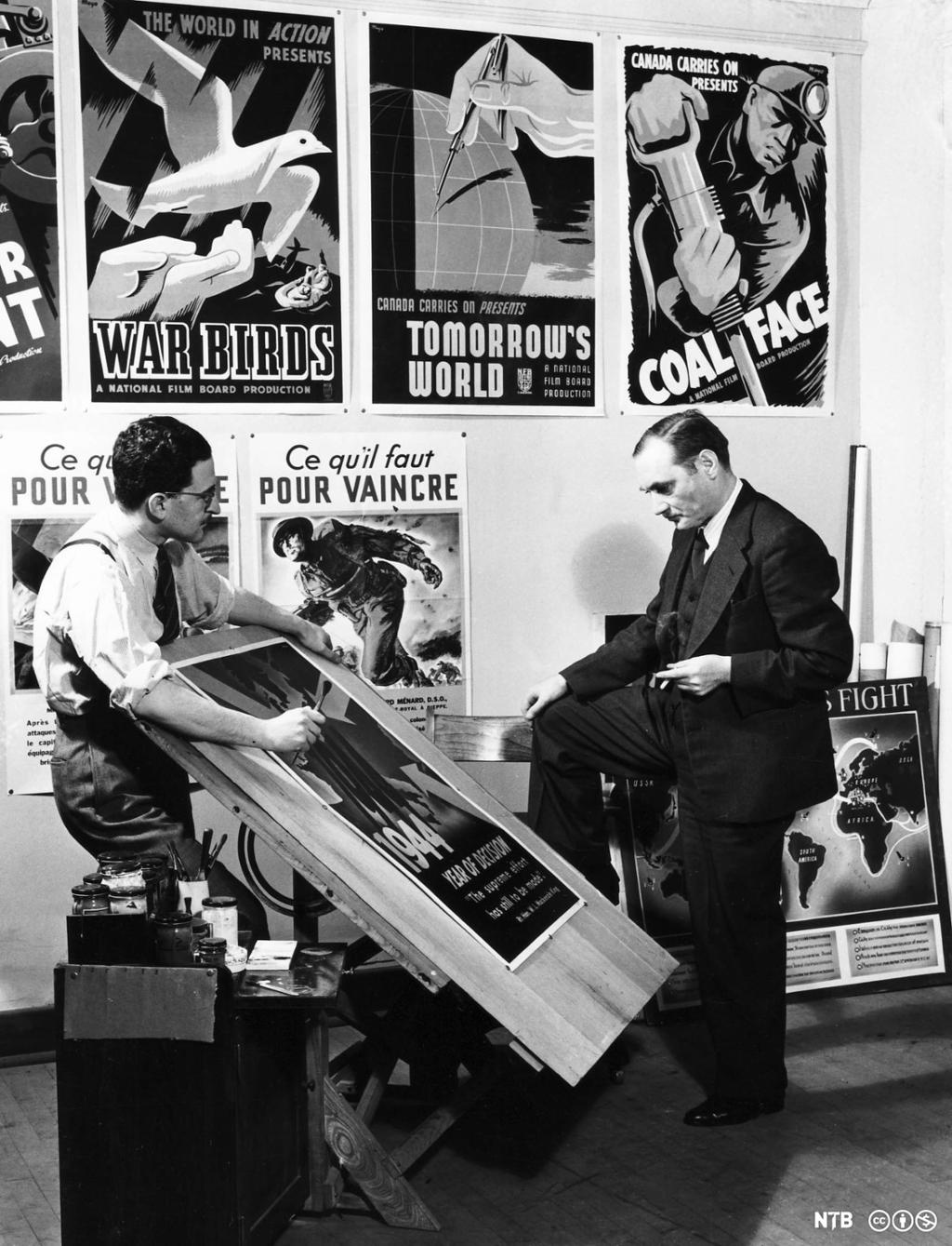 En mann viser fram en plakat, som er montert på et stort tegnebrett, til en annen mann. De står foran en vegg med plakater. Svart-hvitt foto.