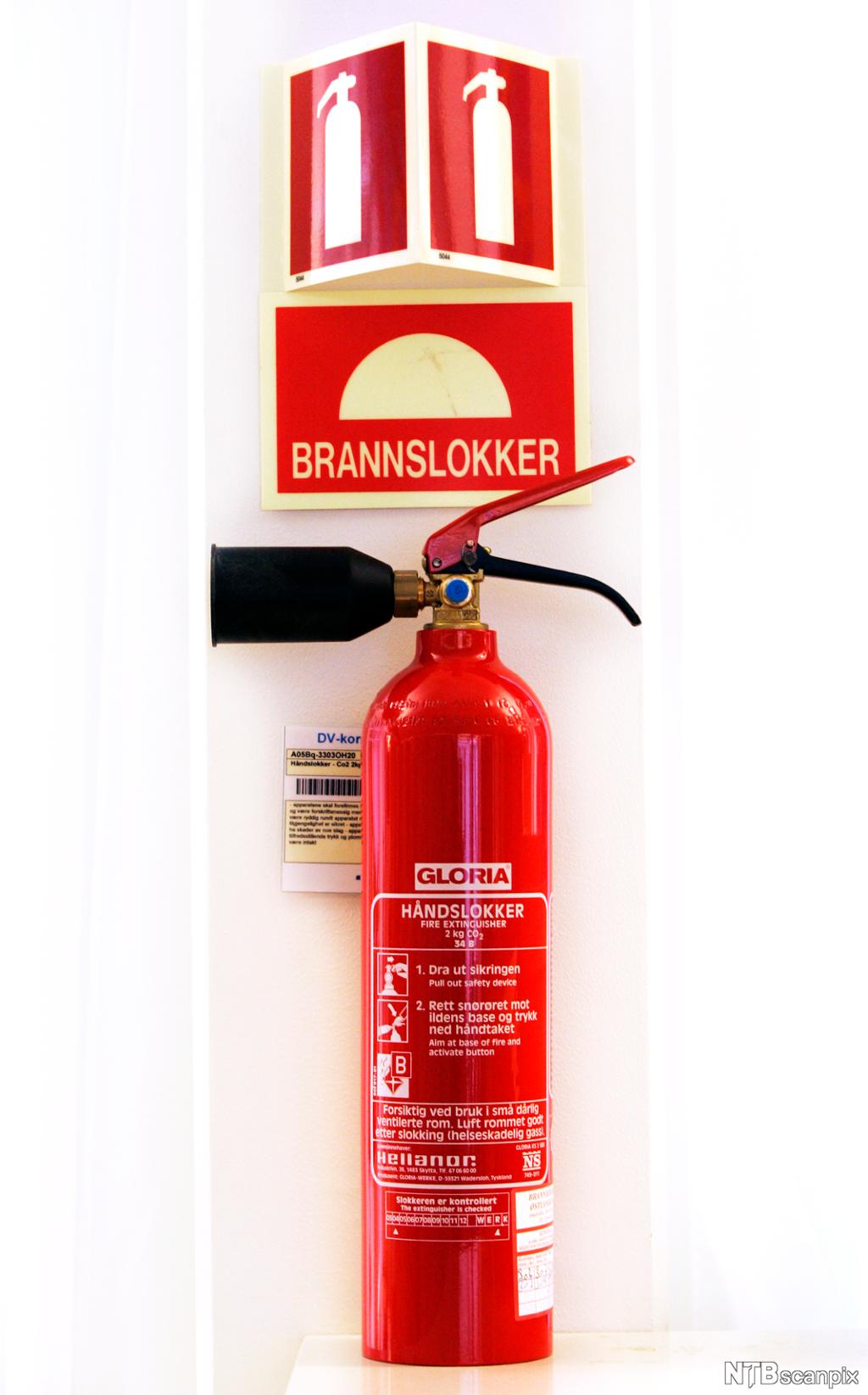 Et rødt håndslukningsapparat. Over apparatet er det ett skilt med en tegning av et brannslukningsapparat, og ett hvor det står "brannslokker". Foto.