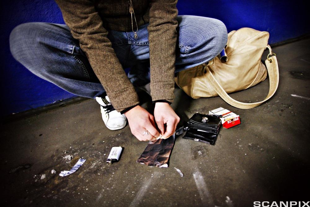 En ung gutt sitter på huk på gata og forbereder stoffet sitt for inntak. Det ligger utstyr som sigarettenner, et stykke aluminiumsfolie, ei lommebok og en pakke sigaretter på asfalten der han bøyer seg ned. Foto.