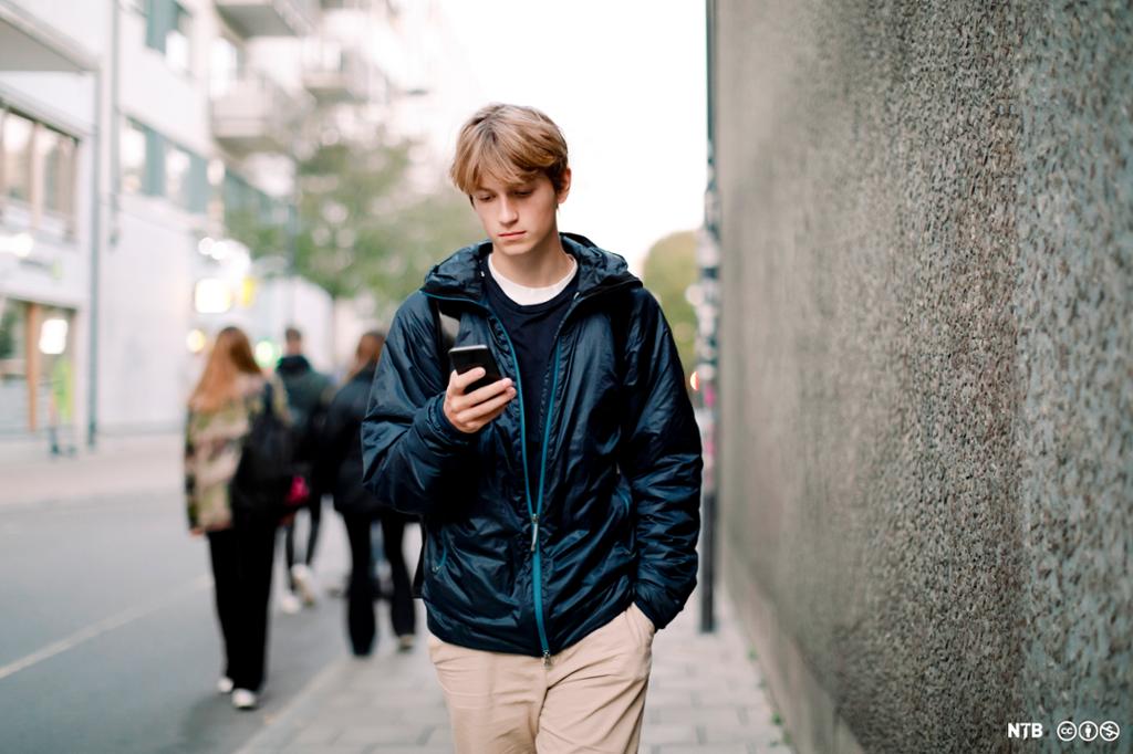 Tenåring går på gata og ser på mobiltelefon. I bakgrunnen er flere personer som går i samme gate. Foto.