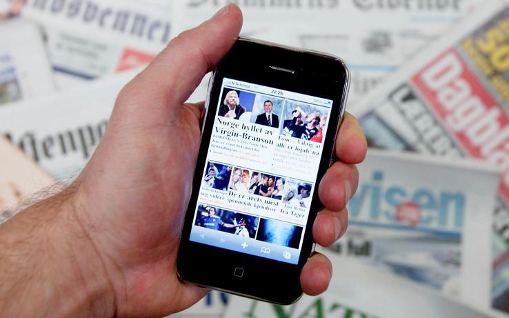 Hånda til en mann holder en smarttelefon med ei nettavis på skjermen. I bakgrunnen ligger forskjellige papiraviser. Foto.