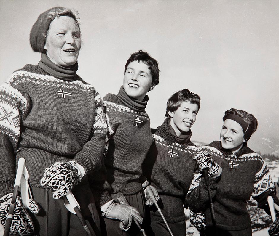 Fire smilende kvinner med strikkegensere med et norsk flagg på brystet. Svart-hvitt foto.