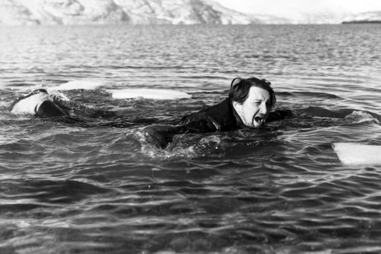 En svartkledd mann svømmer på åpent hav med fjell i horisonten. Svart-hvitt-foto.