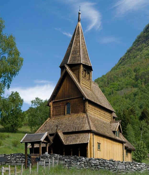 Urnes stavkirke i grønne omgivelser med steinmur foran og fjell bak. Foto.