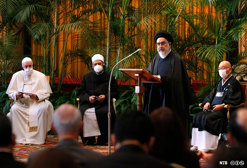En iransk religiøs leder med kappe, skjegg og turban holder en tale ved en talerstol foran andre religiøse ledere som sitter rundt ham i et lokale. Foto.