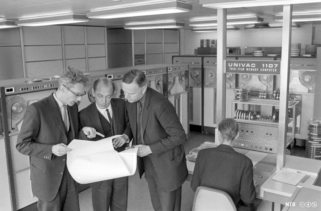 Tre menn i dress studerer et ark med utskrift av programmeringskode. En fjerde mann sitter med ryggen til ved en datamaskinterminal av typen "Univac 1107". Foto.