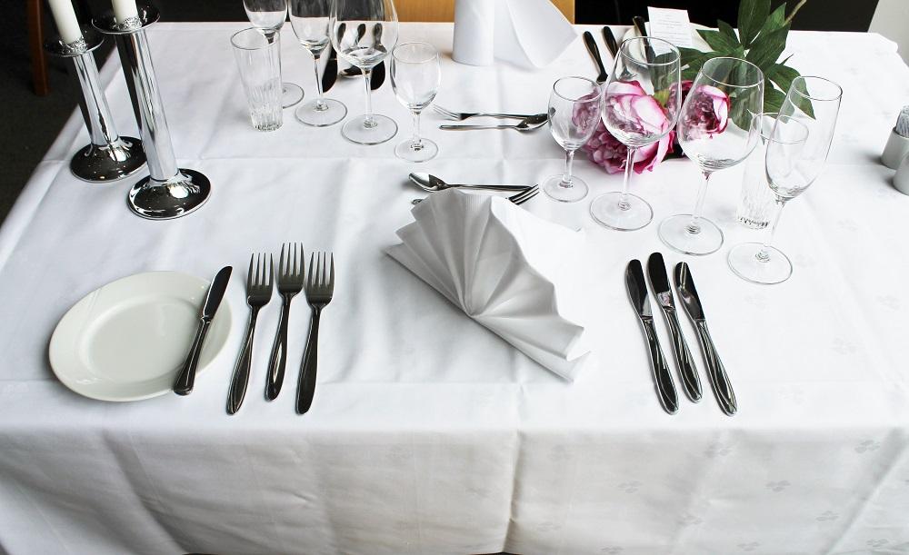 Fint dekket bord med hvit duk, bestikk til flere retter, kuvertasjett, glass, serviett, lysestaker og blomster. Foto.
