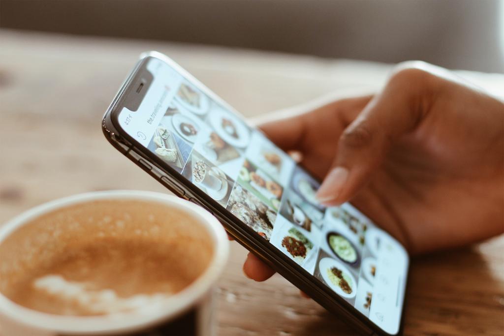 Nærbilde av en kaffekopp og en hånd som holder en mobiltelefon og blar på Instagram. Foto.