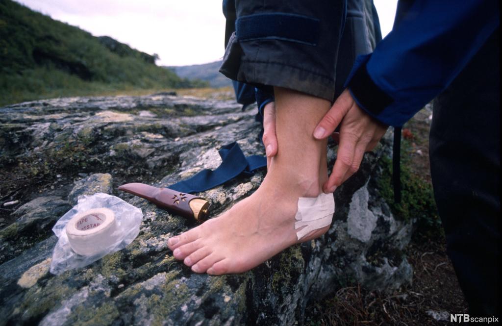 En fot med en teipbit på hælen i naturomgivelser. En sportsteip og en kniv ligger ved siden av. Foto.