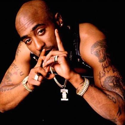 A photo of the rapper Tupac Shakur - 2Pac. He's wearing a lot of bling and has numerous tatoos on his arms. He's looking directly into the camera. 