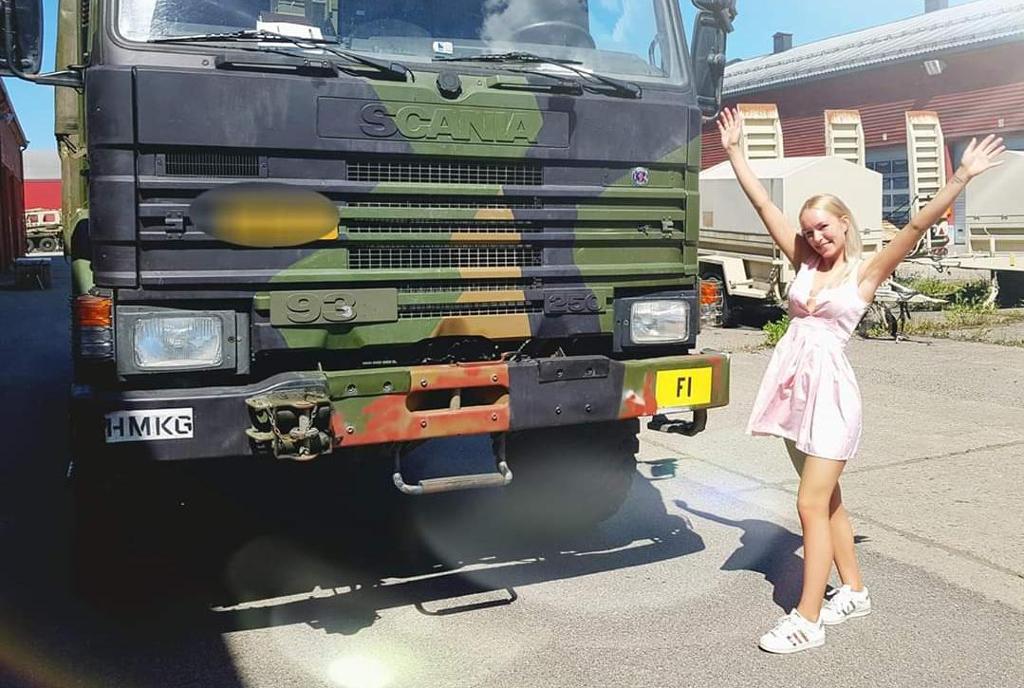Mathilde Grøndalen Lund står ved siden av en stor lastebil. Hun er kledd i rosa kjole og hvite sneakers, og hun holder hendene i været og smiler. Foto.