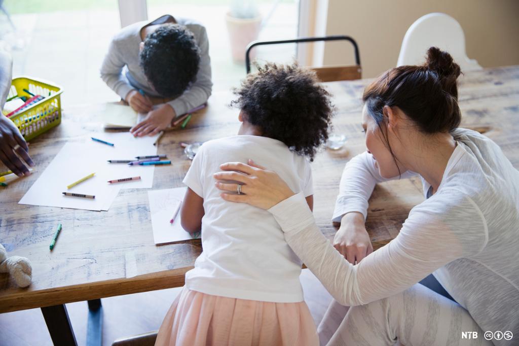 To barn tegner ved et bord, og en voksen kvinne sitter ved siden av og holder en hånd på ryggen til det ene barnet. Foto.  