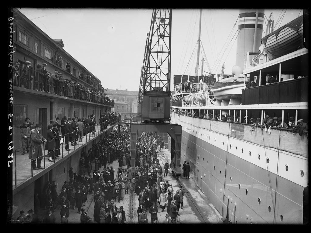 Et stort skip ligger ved kai. Mange folk står ved rekka på skipet, og mange står på kaia. Ei kran står på kaikanten. Svart-hvitt foto. 