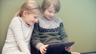 Liten gutt og jente sitter på gulvet og ser på en iPad sammen. De smiler. Foto.