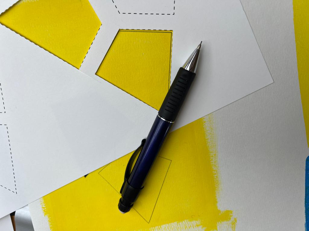 Måla flate med gulfarge på eit ark. Over ligg ein sjablong og ein blyant. Det er teikna opp former etter sjablongen på den måla flata. Foto.
