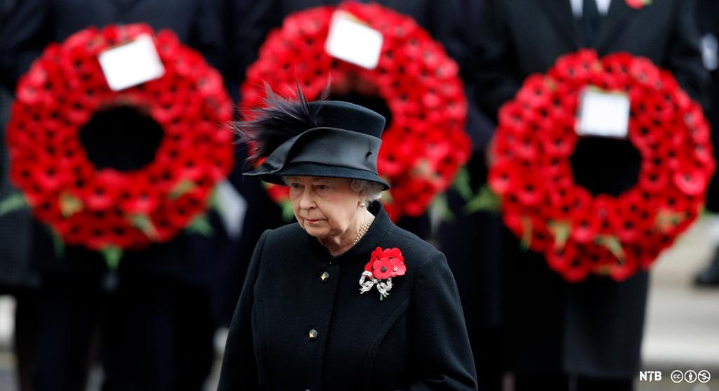 Photo of Queen Elizabeth II; she is an old woman dressed in black. She is wearing a bouquet of artificial poppies on her chest. In the background, there are three poppy wreaths displayed. 