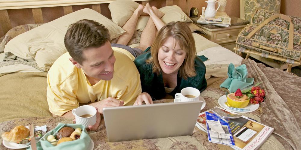 Eit par bestiller noko på ein berbar PC mens dei et frukost på senga. Foto.