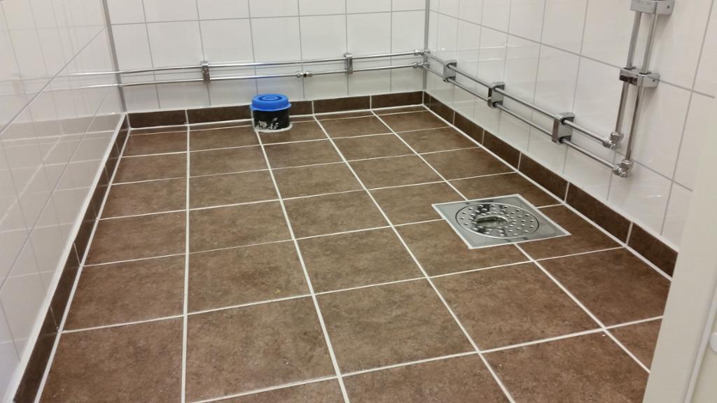 Flislagt gulv med sluk og nedre del av vegg med baderomspanel og synlige rør for vann. Foto.