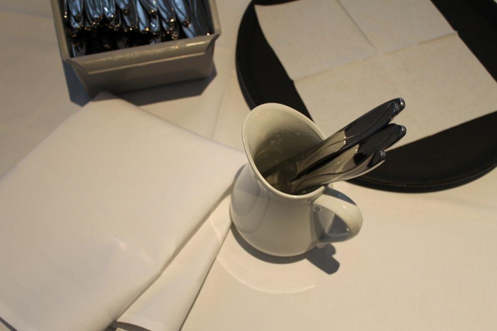 En boks med bestikk, et serveringsbrett med en serviett på, en mugge med vann  hvor det står kniver oppi, og en tøyserviett liggende på et bord. Foto.