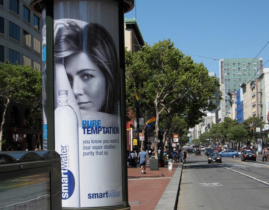 Et urbant miljø der det er montert en reklameplakat på en stolpe langs en trafikkert vei. Plakaten reklamerer for flaskevannet smartwater og viser en kvinne og ei flaske fra produsenten, i tillegg til slagord for produktet.  Foto.