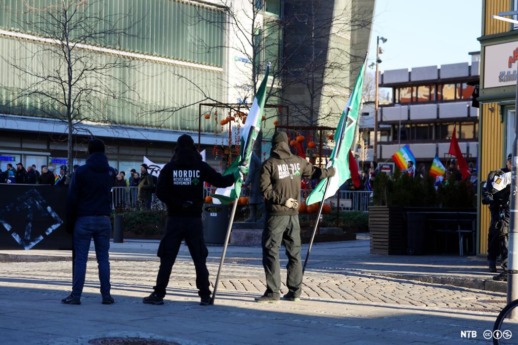 Tre menn med hettegenserar med påskrifta Nordic Resistance Movement står breibeinte på eit torg med ryggen til og held to flagg med organisasjonens logo. I bakgrunnen er det ein stor folkemengde med plakatar og prideflagg. Foto.